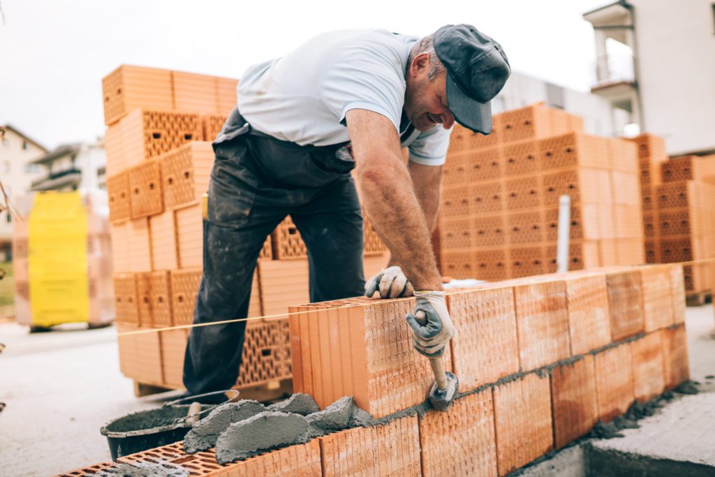 An Image Of A Man Building Exterior Walls, Using Hammer For Laying Bricks In Cement.