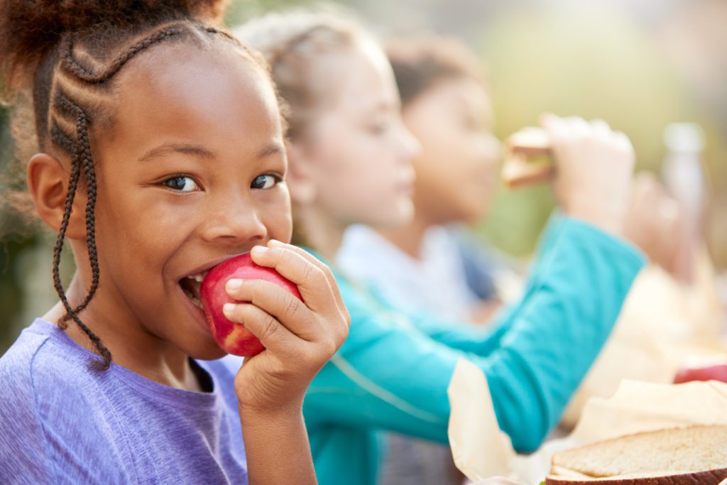 Portrait Of A Little Girl Eating Apple With Her Group of Friends At The Back.
